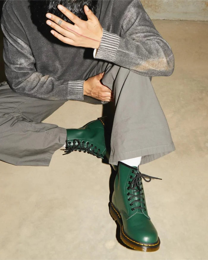 A person seated wearing gray pants and green Dr. Martens 1460 boots with laces.