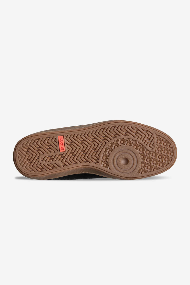 A close-up of a brown rubber shoe sole with a textured herringbone pattern and a small red brand tag, shown against a light gray background.