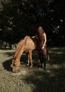 Person in a brown dress standing next to a horse in a grassy field with trees in the background