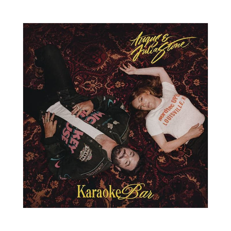 Album cover art for 'Karaoke Bar' by Aqyu & Julia Stone, featuring a top-down photograph of the two artists lying on a dark red patterned oriental rug. Aqyu is wearing a black leather jacket with pink and white graphic patches, and Julia is wearing a white t-shirt with red text.