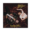 Album cover art for 'Karaoke Bar' by Aqyu & Julia Stone, featuring a top-down photograph of the two artists lying on a dark red patterned oriental rug. Aqyu is wearing a black leather jacket with pink and white graphic patches, and Julia is wearing a white t-shirt with red text.