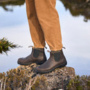Brown Chelsea boots worn with tan pants on a rocky surface with water and greenery in the background