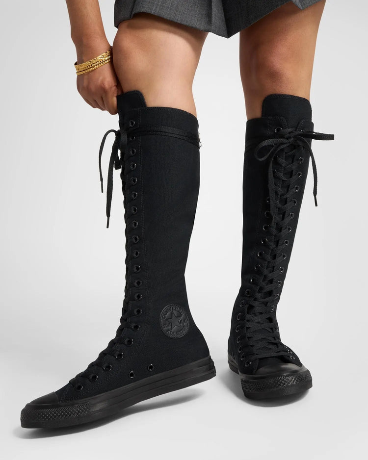 A pair of black knee-high Converse Chuck Taylor All Star Foundation XX-Hi sneakers with lace-up front and a zipper at the back, worn by a person standing.