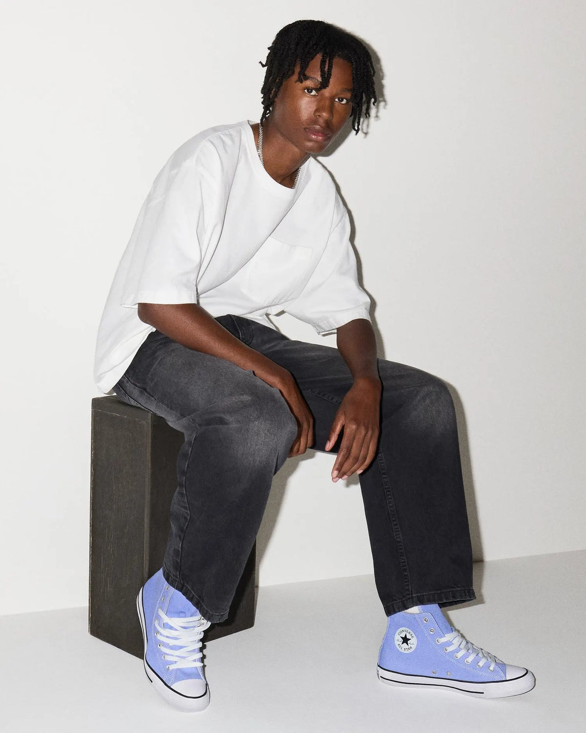Person wearing a white oversized shirt, black jeans, and blue Converse sneakers sitting on a block.
