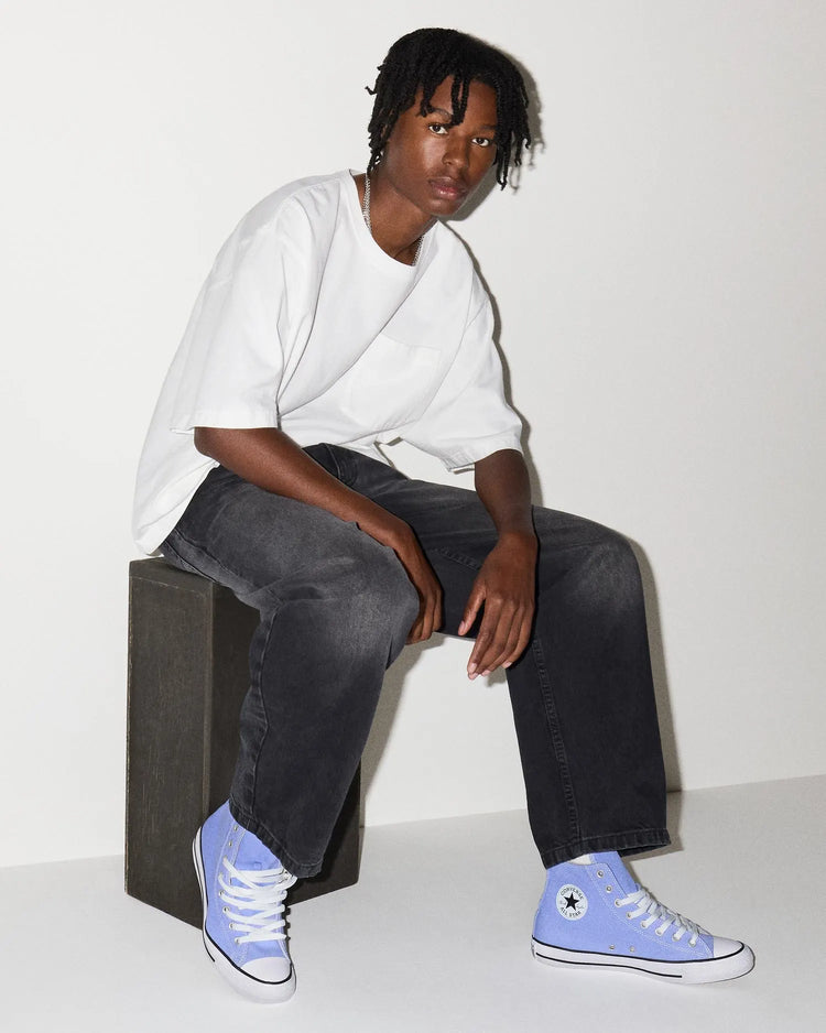 Person wearing a white oversized shirt, black jeans, and blue Converse sneakers sitting on a block.