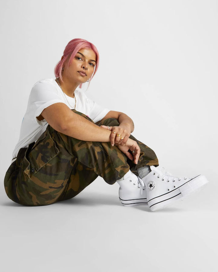 A person seated wearing white high-top Converse Chuck Taylor All Star Lift Leather sneakers with a camouflage pattern.