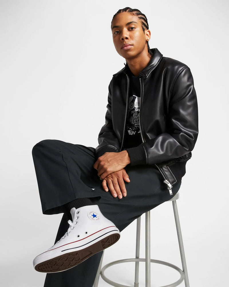 Person wearing a black leather jacket and black pants sitting on a stool against a white background