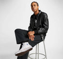 Person wearing a black leather jacket and white sneakers sitting on a stool against a plain background