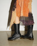 A person wearing black lace-up ankle boots with a patterned orange, purple, and beige skirt, standing against a plain light wall.