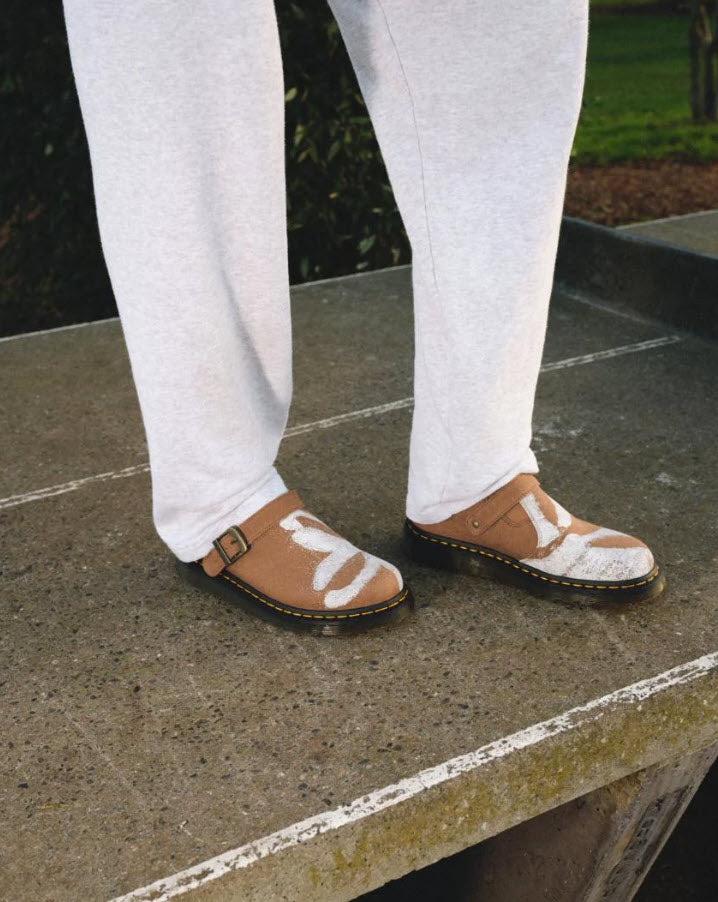 A person wearing white trousers and brown hemp canvas vegan mules with a floral print, standing on a concrete surface.