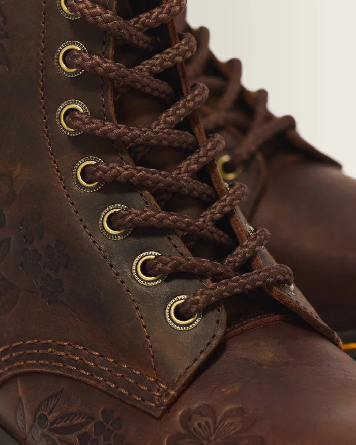 Close-up of a dark brown leather lace-up boot with braided laces, brass eyelets, and floral embossing on the side.