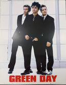 Green Day band poster featuring the three members in black suits standing in front of a bright white paneled wall, with the band name 'GREEN DAY' in large red letters at the bottom.