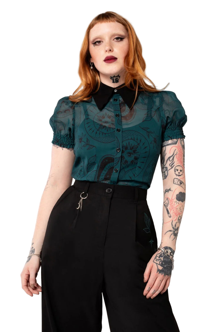 Person wearing a teal blouse with black collar and black pants on a light pink background