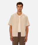 Man wearing a beige short-sleeve shirt over a white shirt on a plain background