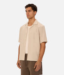 Man wearing a beige short-sleeve shirt over a white shirt on a plain background