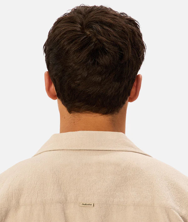 Back view of a person wearing a beige shirt with a visible brand logo on a light background