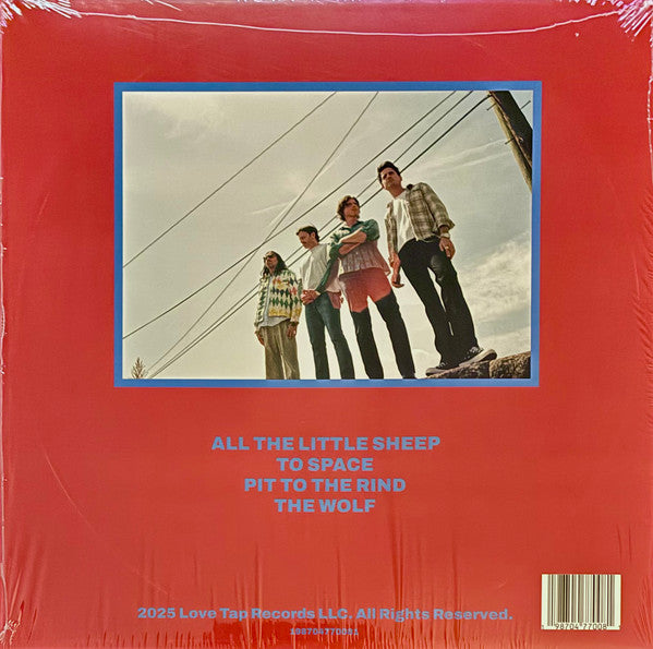 The back cover of a vinyl record album, sealed in plastic shrink wrap. The cover is a solid red color with a blue-bordered rectangular photo of four men standing outdoors under power lines.
