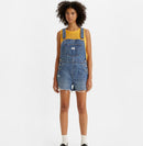 A woman standing, wearing a pair of vintage-style blue denim shortalls with adjustable straps and multiple pockets.