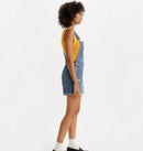 A woman standing, wearing a pair of vintage-style blue denim shortalls with adjustable straps and multiple pockets.