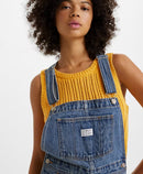 A woman standing, wearing a pair of vintage-style blue denim shortalls with adjustable straps and multiple pockets.