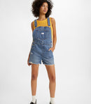 A woman standing, wearing a pair of vintage-style blue denim shortalls with adjustable straps and multiple pockets.