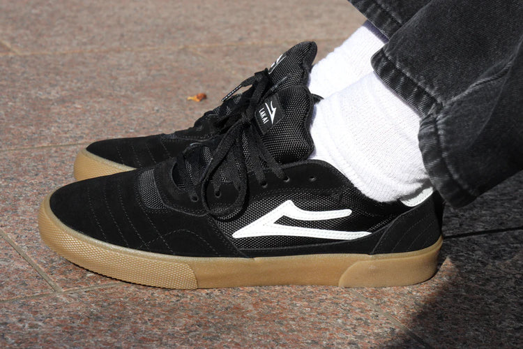 Black sneakers with tan soles worn by a person on a pavement.