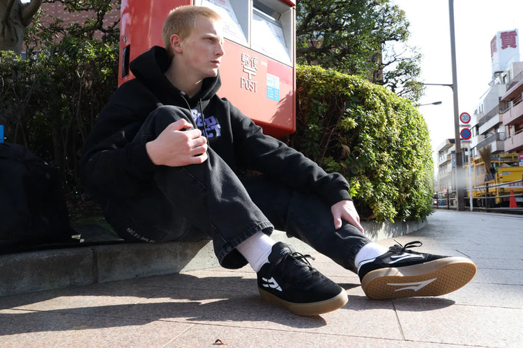 Person sitting on a sidewalk wearing black sneakers with white soles.