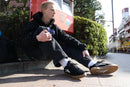 Person sitting on a sidewalk wearing black sneakers with white soles.