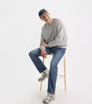 Person sitting on a stool wearing a gray sweater, blue jeans, and a cap on a white background