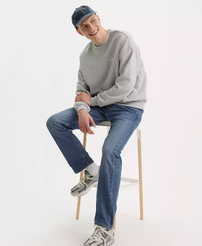 Person sitting on a stool wearing a gray sweatshirt, blue jeans, and a cap on a white background