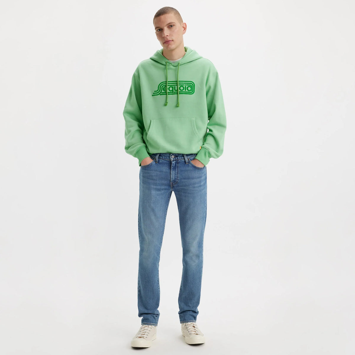 Person wearing a green hoodie with a logo and blue jeans on a white background