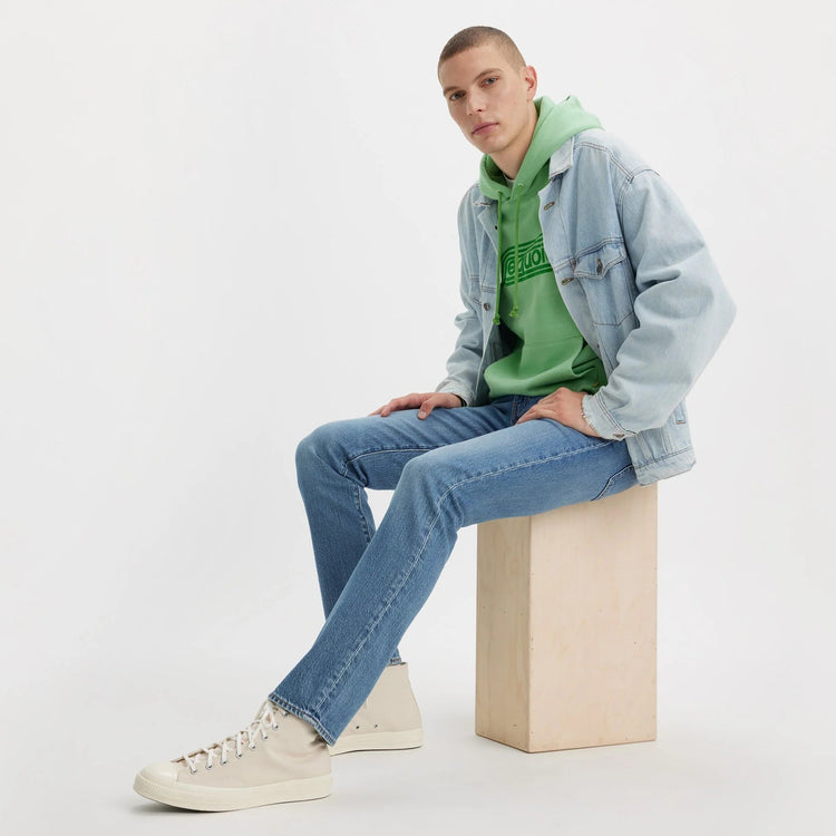 Person sitting on a block of wood wearing a green hoodie, denim jacket, and jeans against a white background