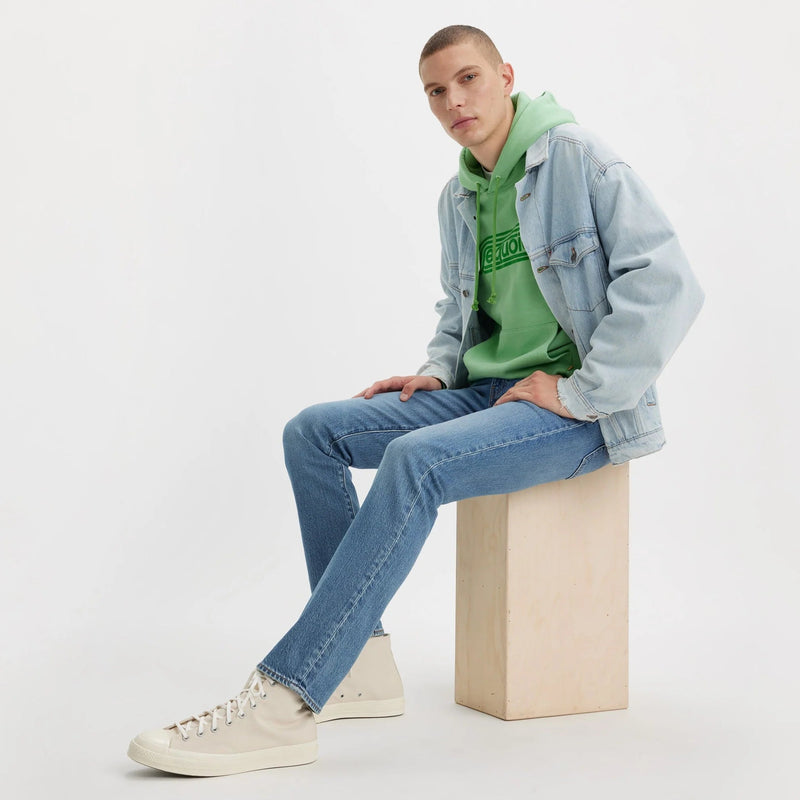 Person sitting on a block of wood wearing a green hoodie, denim jacket, and jeans against a white background