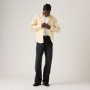 Man wearing a beige shirt over a white t-shirt with black pants on a light gray background