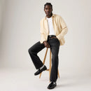 Man sitting on a stool wearing a beige jacket, white shirt, black pants, and black shoes on a plain background