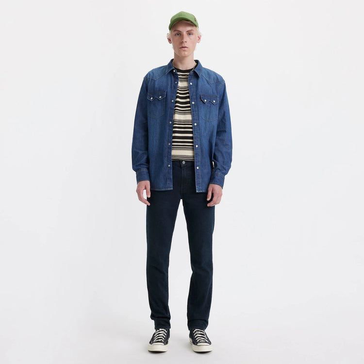 Man wearing a blue denim jacket, striped shirt, and dark jeans on a white background