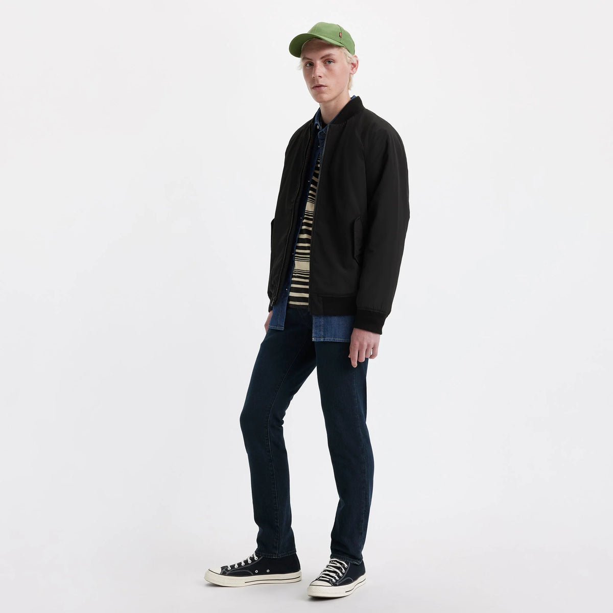 Person wearing a black jacket, striped shirt, jeans, and green cap on a white background