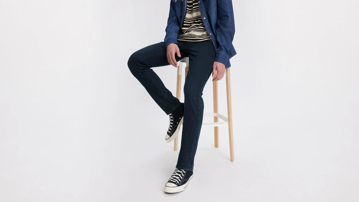 Person wearing dark jeans and a blue jacket sitting on a stool against a white background