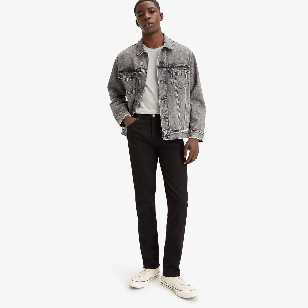 Man wearing a gray denim jacket, white shirt, black pants, and white sneakers on a white background