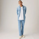 Man wearing a light blue denim shirt over a white t-shirt, paired with light blue jeans and white sneakers on a plain background.
