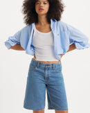 Woman wearing a light blue shirt jacket, white tank top, and blue shorts on a white background