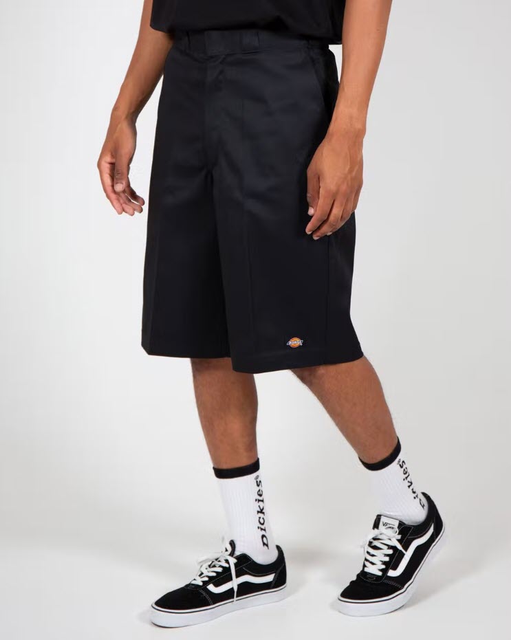 A person standing wearing Dickies black multi-pocket work shorts with a loose fit and knee-skimming hem, paired with white socks and black sneakers.
