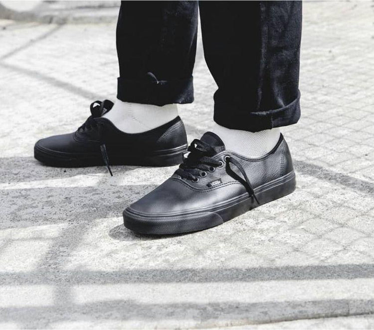 A pair of black Vans Authentic leather sneakers with lace-up closure, displayed on a cobblestone surface.